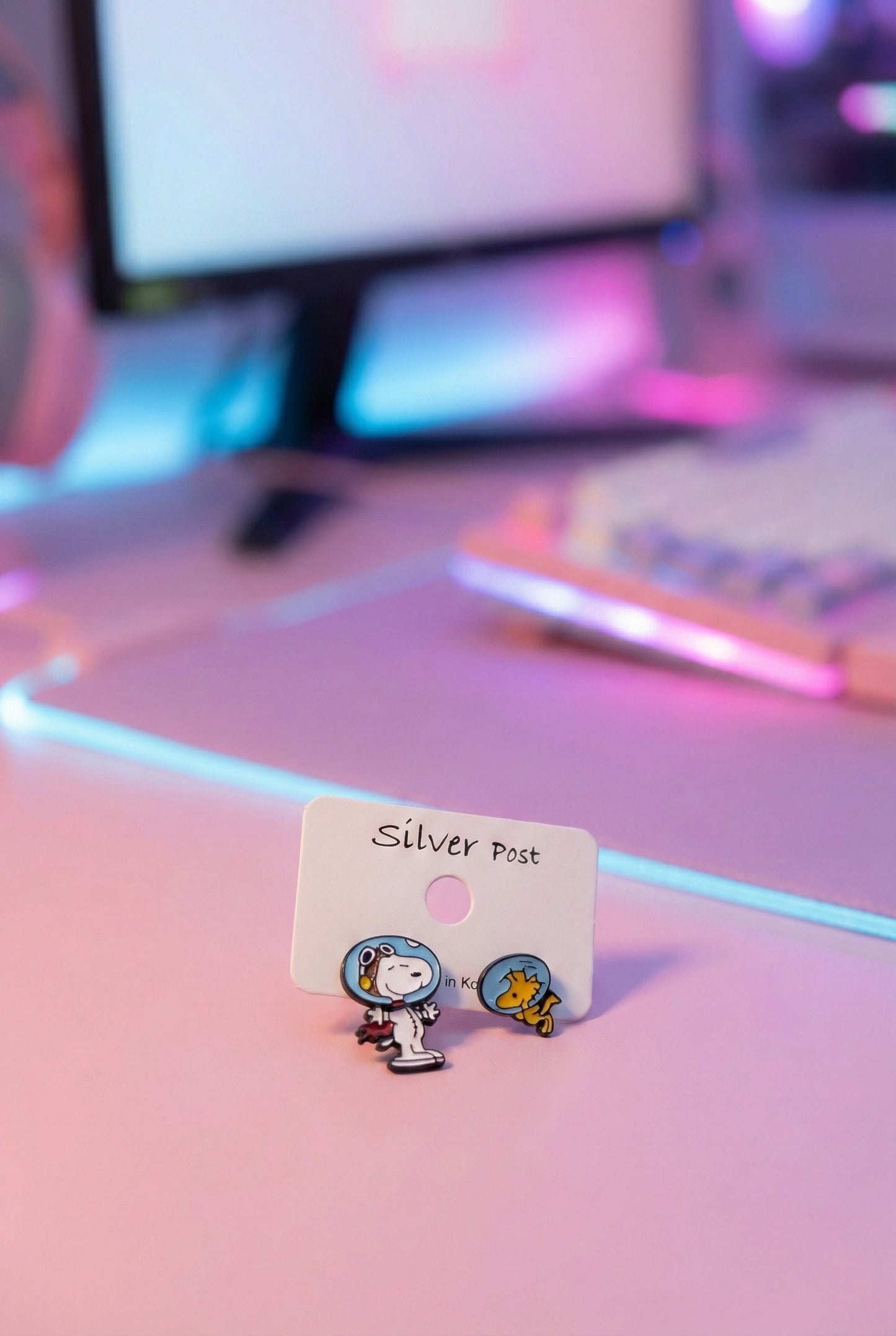 Snoopy and Woodstock astronaut earrings displayed on a white earring card marked “Silver Post,” with a pink-purple gradient computer desk background.