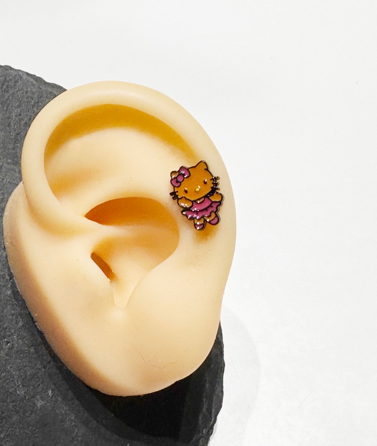 Adorable Hello Kitty–inspired piercing featuring a playful Hawaii-style cat in pink or red dress. Made with hypoallergenic surgical steel. Sold as single.