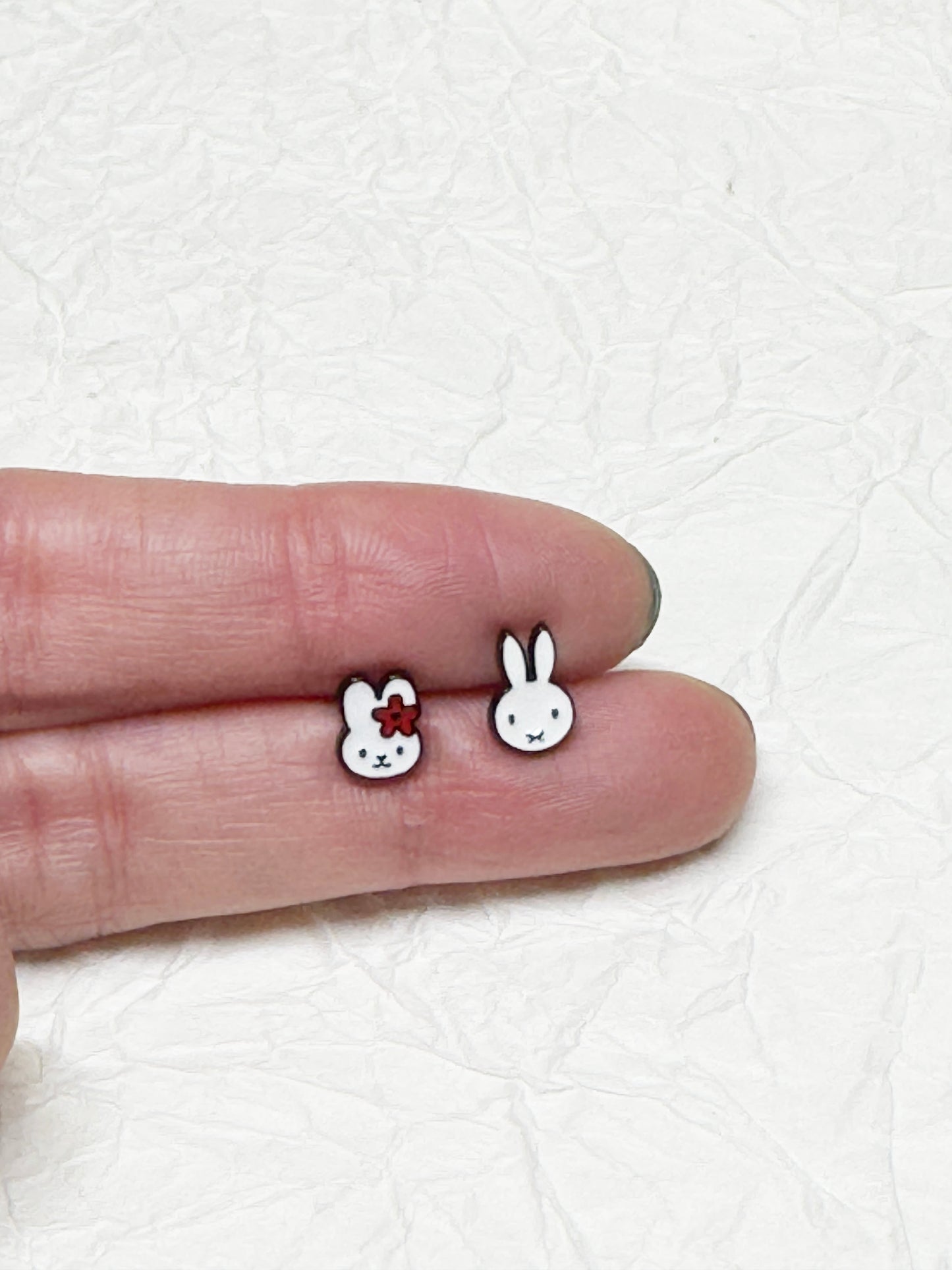 Miffy character earrings shown on fingertips for size comparison; one has a red flower, the other is a classic outline — placed over a textured white paper background.