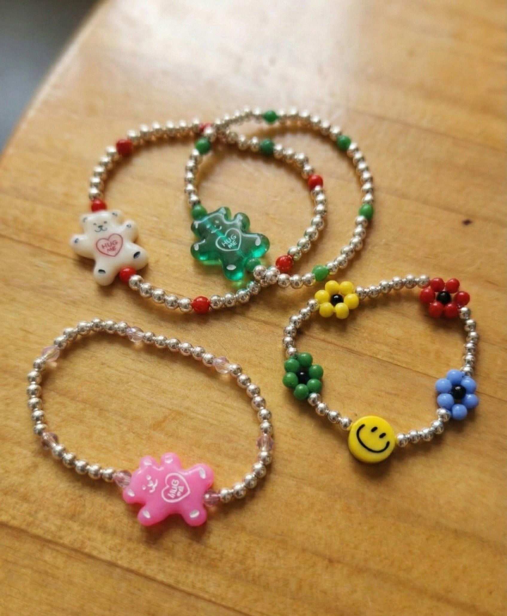 Four 925 sterling silver beaded bracelets flat lay: pink gummy bear, white gummy bear, green gummy bear, and smiley garden flower bracelet on wooden surface
