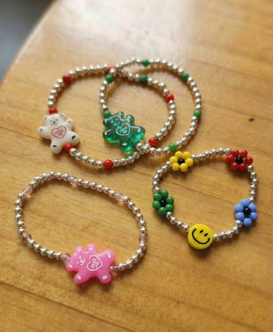Four 925 sterling silver beaded bracelets flat lay: pink gummy bear, white gummy bear, green gummy bear, and smiley garden flower bracelet on wooden surface
