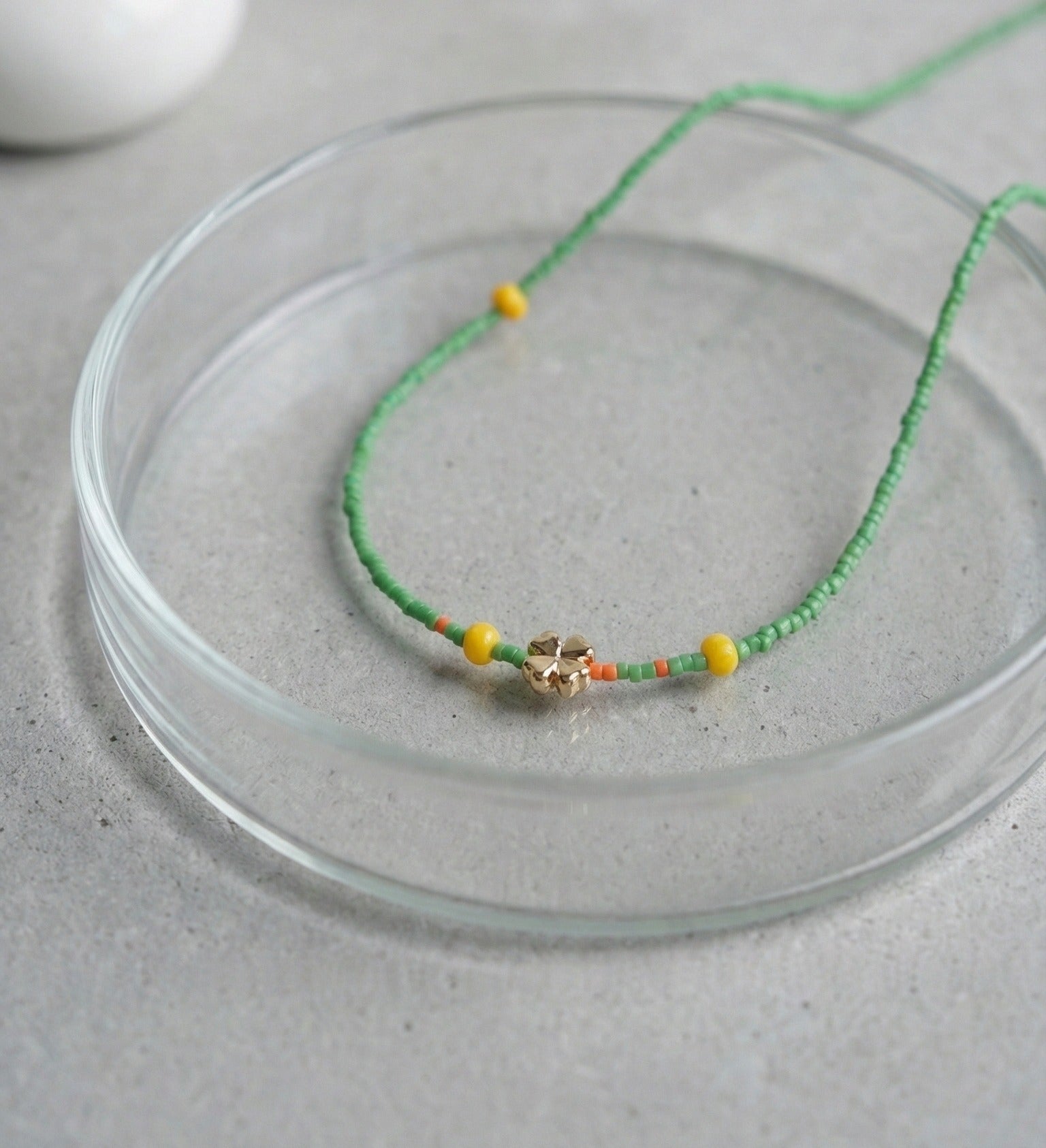 Green seed bead necklace with gold four-leaf clover charm and yellow accent beads displayed on glass plate