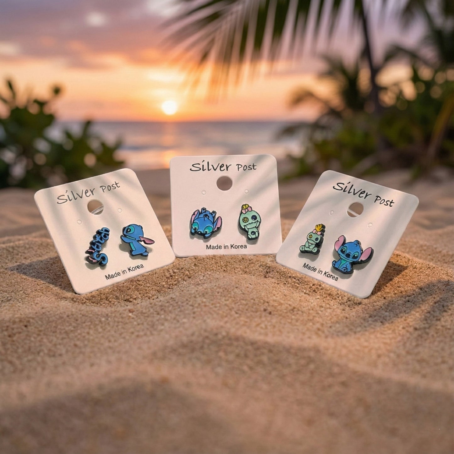 Three pairs of Stitch and Scrump earrings on display cards in the sand at sunset, silver post, Made in Korea.