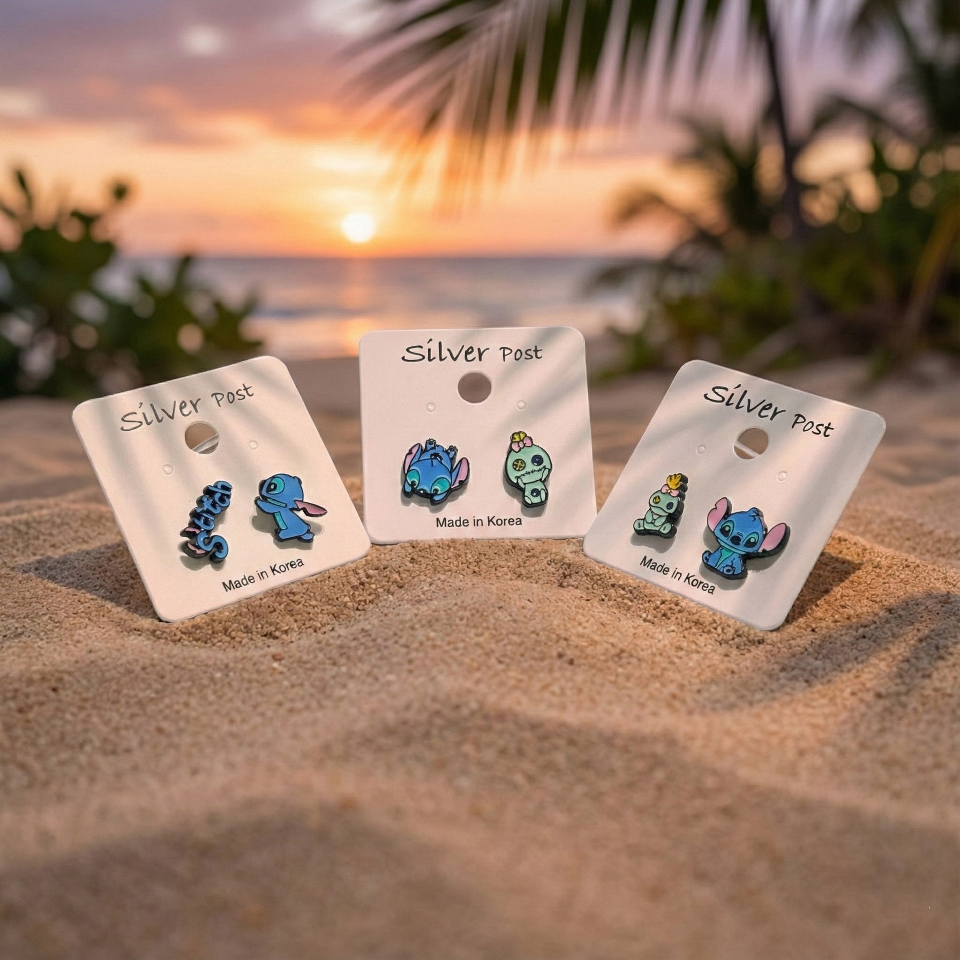 Three pairs of Stitch and Scrump earrings on display cards in the sand at sunset, silver post, Made in Korea.