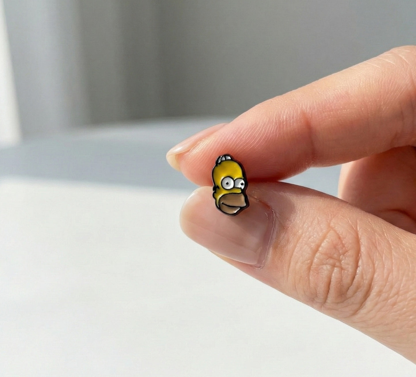Homer Simpson mini enamel stud earring held between fingers, showing yellow character face detail