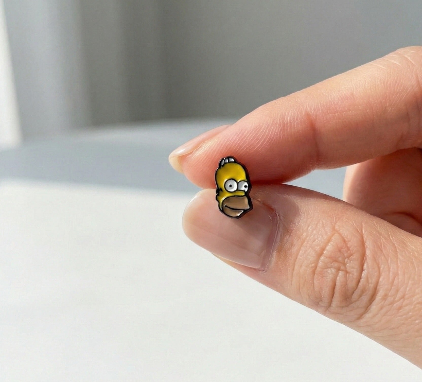 Homer Simpson mini enamel stud earring held between fingers, showing yellow character face detail