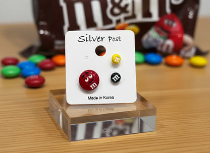M&M's character earring set displayed on white card with silver post label on clear acrylic stand, featuring red smiling face, yellow and black mini studs, colorful M&M's candies scattered in background, made in Korea packaging