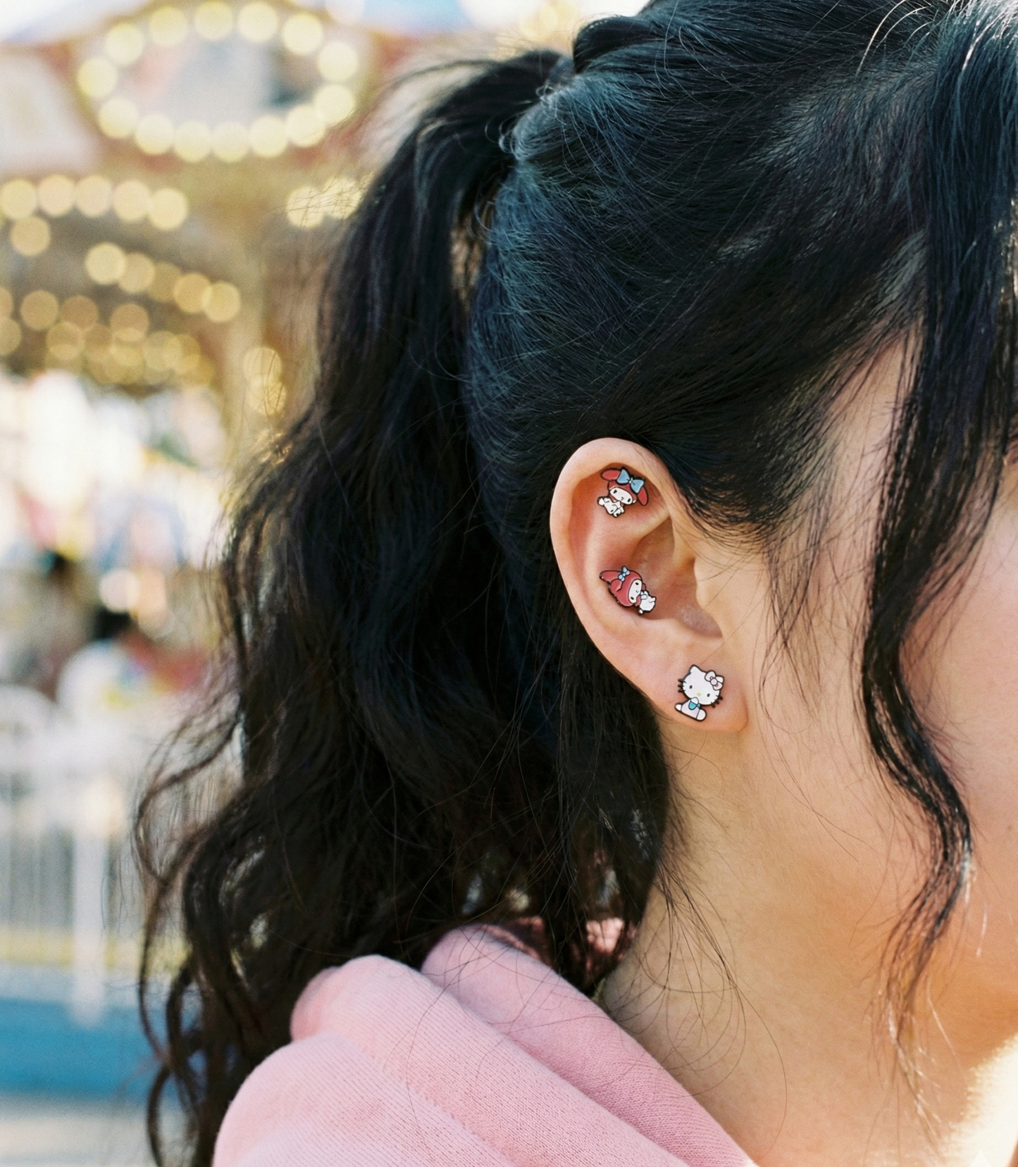 Side view of ear wearing My Melody helix piercing and Kuromi lobe stud at amusement park