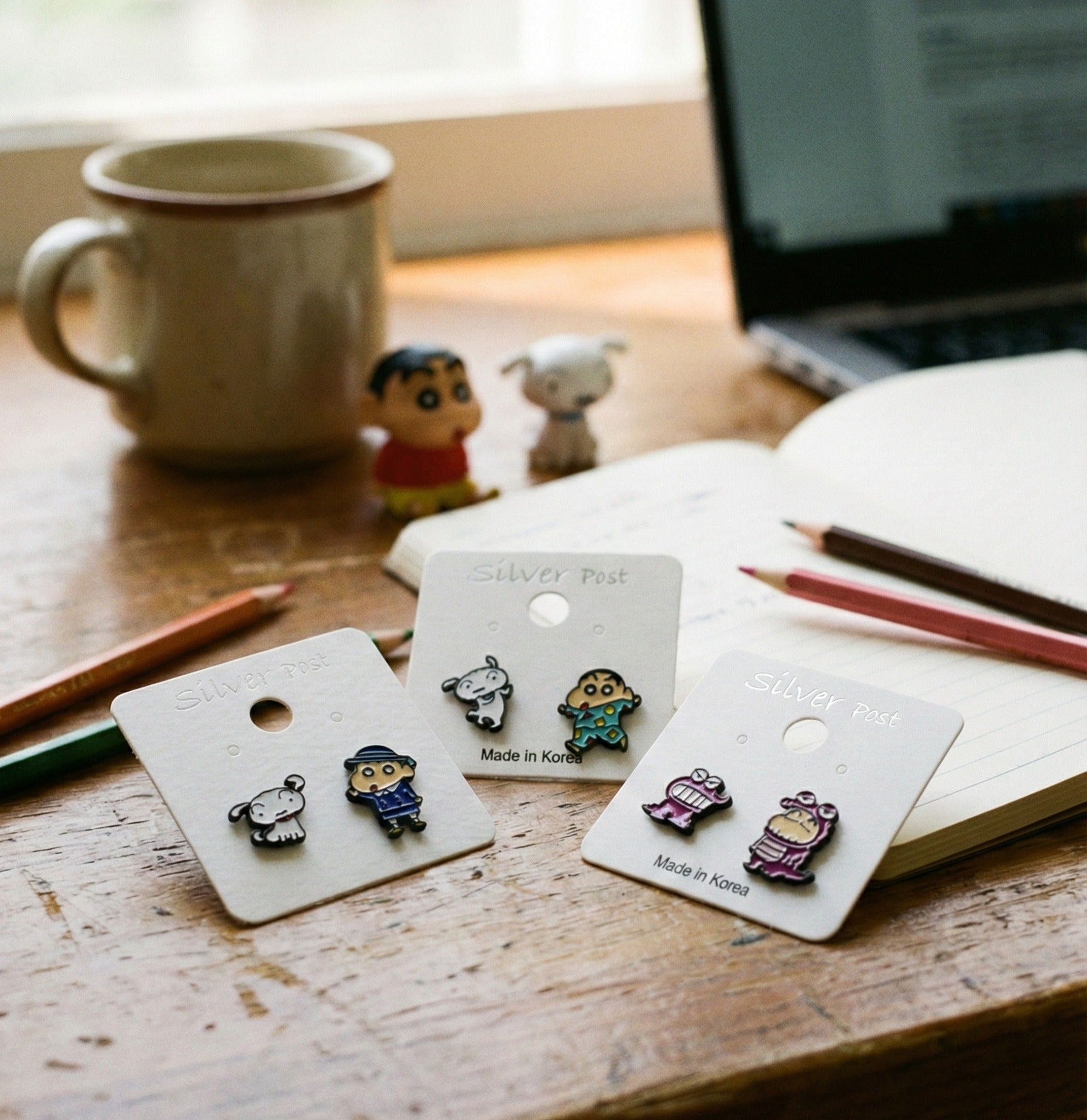 Lifestyle flatlay of Shin-chan earrings on wooden desk with notebook, pencils, and coffee mug