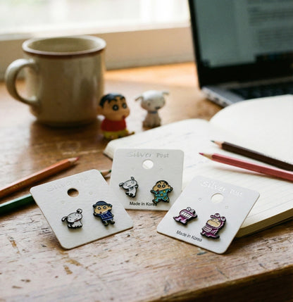 Lifestyle flatlay of Shin-chan earrings on wooden desk with notebook, pencils, and coffee mug