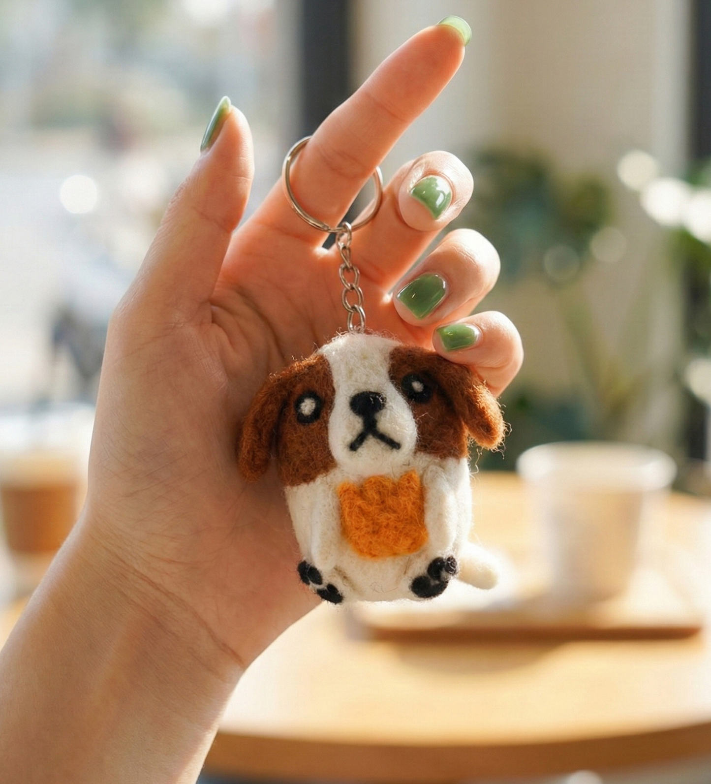 Hand holding handmade wool felt dog keychain with heart detail in cafe setting with green nail polish
