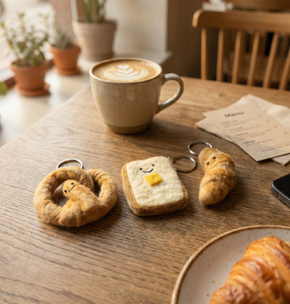 Three handmade bakery felt keychains - pretzel, toast and croissant on wooden cafe table