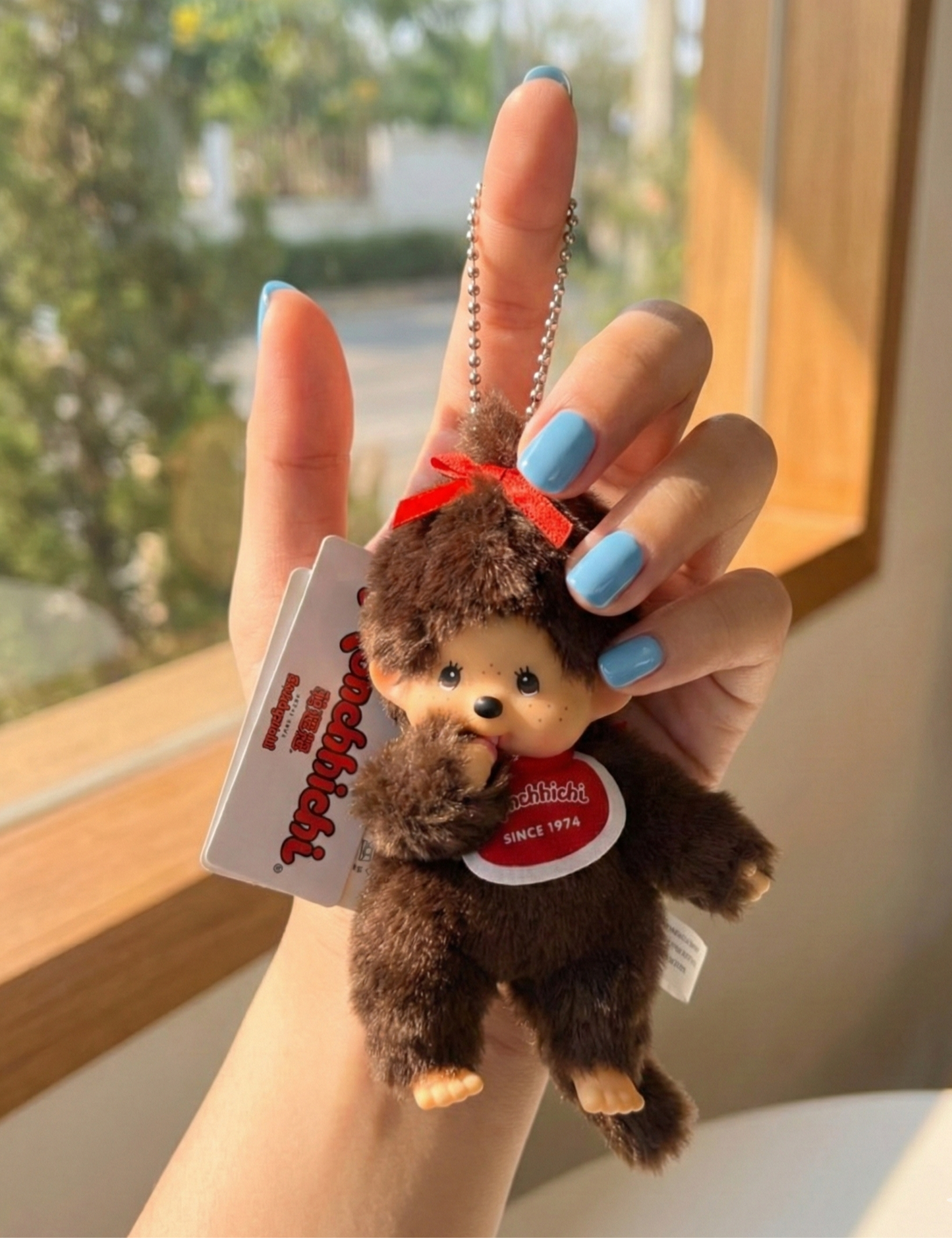 Hand holding Monchhichi Ribbon Girl plush keychain with red bow near sunny window — kawaii Japanese character bag charm