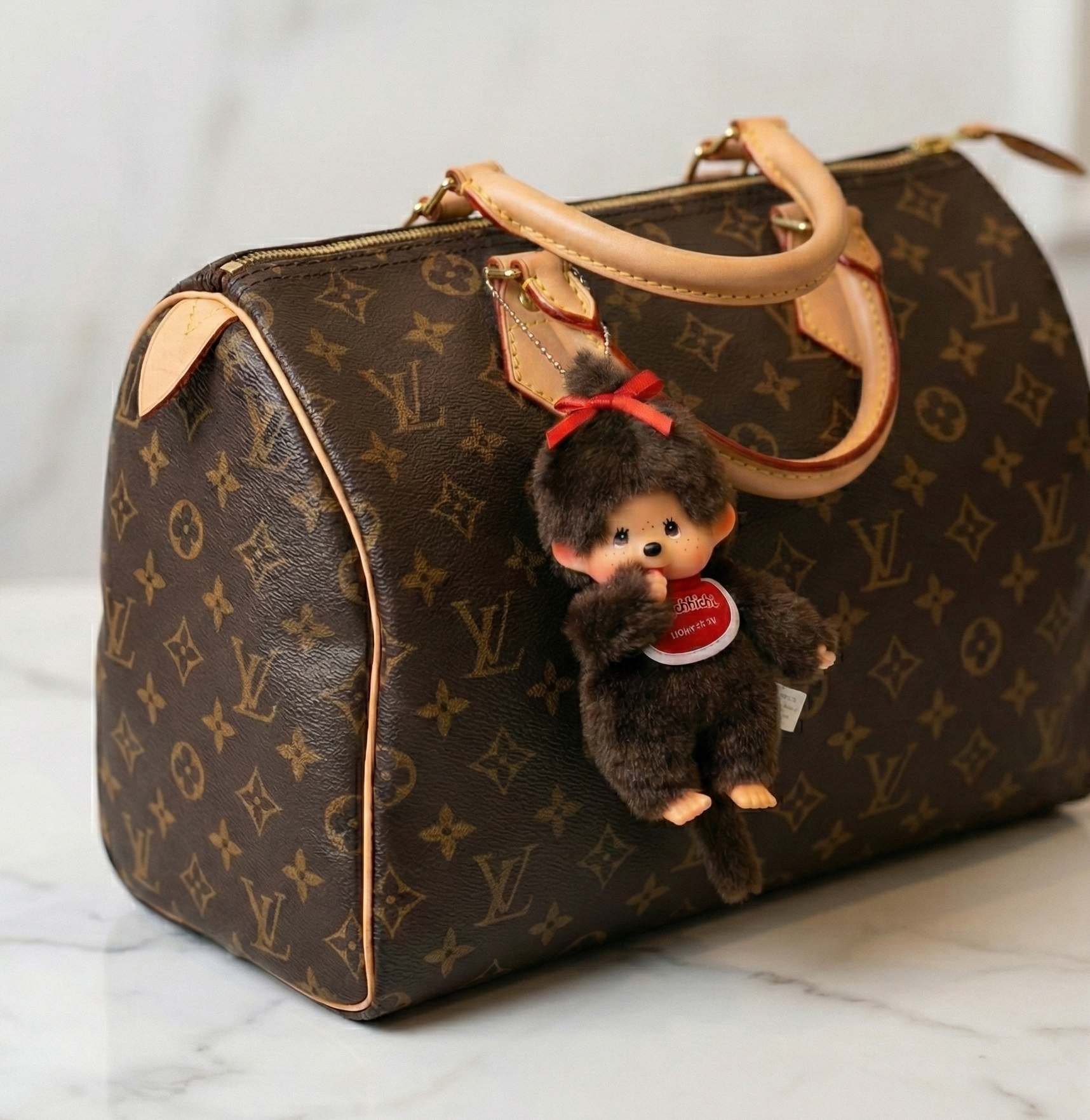  Monchhichi Ribbon Girl plush bag charm hanging from Louis Vuitton Speedy monogram bag on marble surface — luxury bag charm styling