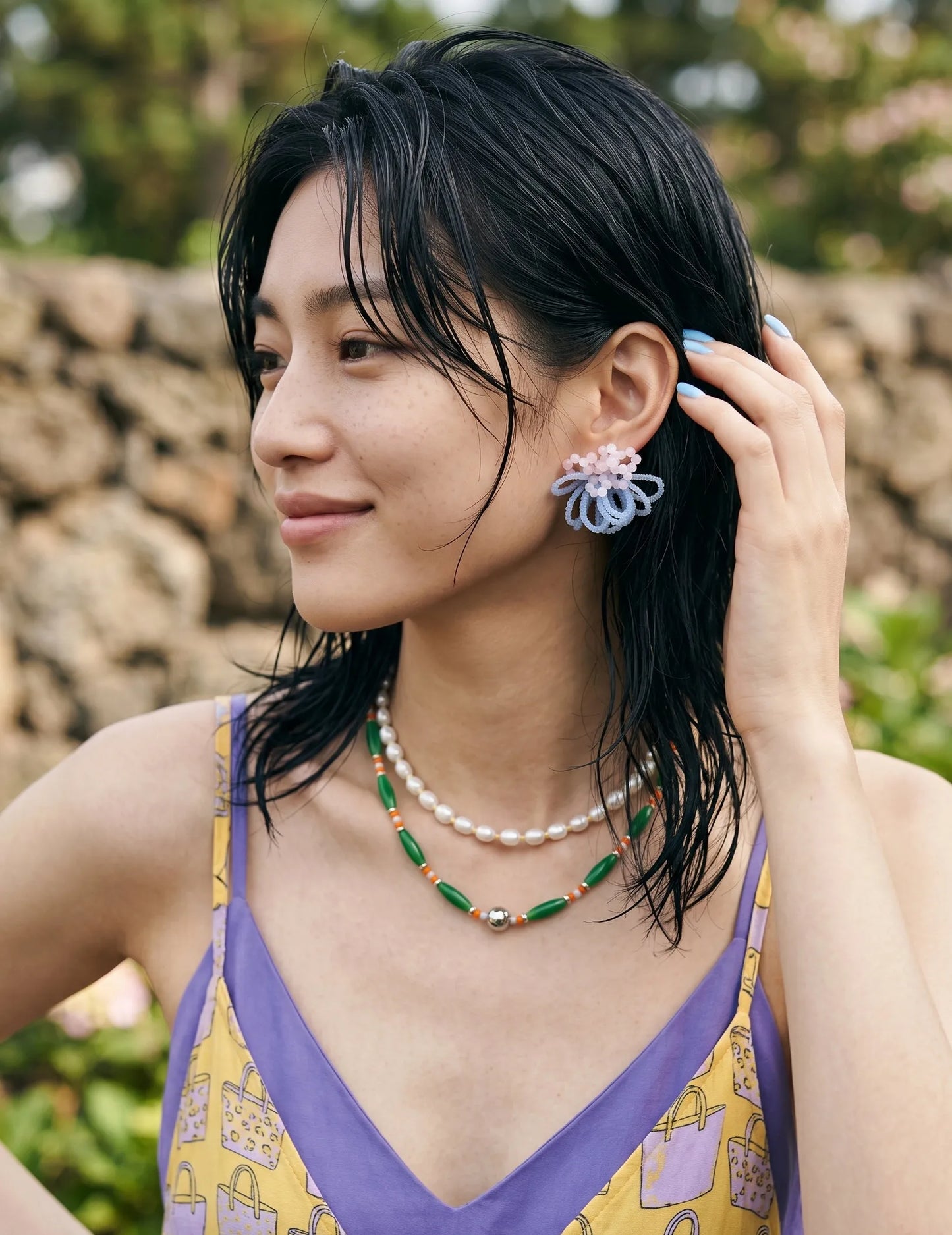 A portrait of a woman with trendy wet-look hair wearing handcrafted pink and light blue beaded cluster earrings. She styles the statement jewelry with a Y2K-inspired layered pearl and green bead necklace over a colorful summer slip dress against a natural stone wall.