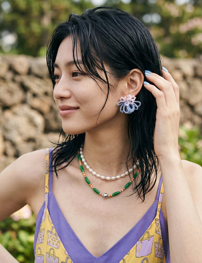 A portrait of a woman with trendy wet-look hair wearing handcrafted pink and light blue beaded cluster earrings. She styles the statement jewelry with a Y2K-inspired layered pearl and green bead necklace over a colorful summer slip dress against a natural stone wall.