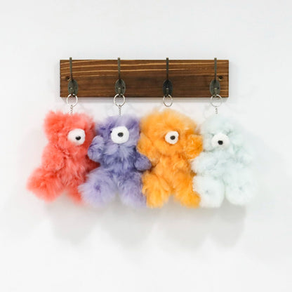 Four alpaca bear bag charms in coral, purple, orange and mint hanging from a rustic wooden wall hook rack against a white background
