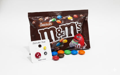 M&M's character earrings displayed with chocolate candy bag on white surface, product card showing silver post detail and Made in Korea label, playful candy-themed jewelry styling with scattered colorful M&M's chocolates