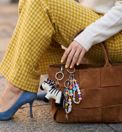 Zebra felt keychain with colorful beads attached to woven brown tote bag with yellow gingham coat in background