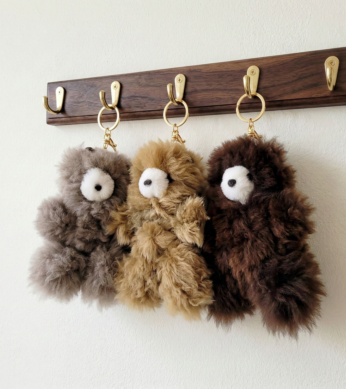 Three alpaca fur bear keyrings in gray, caramel-beige and dark chocolate brown hanging from a wall-mounted wooden rack with gold hooks
