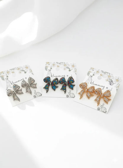 Three colorways of crystal bow stud earrings on jewelry cards — Antique Multi (dark with multicolor crystals), Silver Blue (antique silver with blue crystals), and Gold Pink (gold with pink crystals) — handmade in Korea
