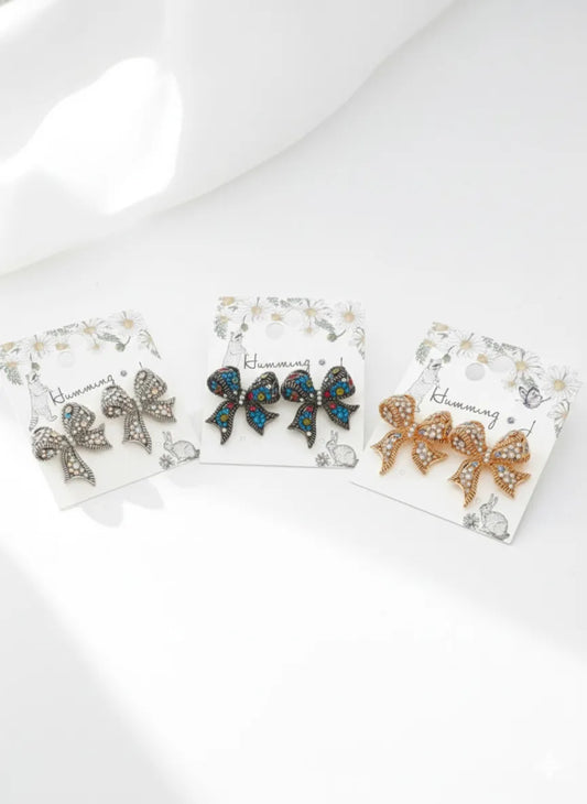 Three colorways of crystal bow stud earrings on jewelry cards — Antique Multi (dark with multicolor crystals), Silver Blue (antique silver with blue crystals), and Gold Pink (gold with pink crystals) — handmade in Korea