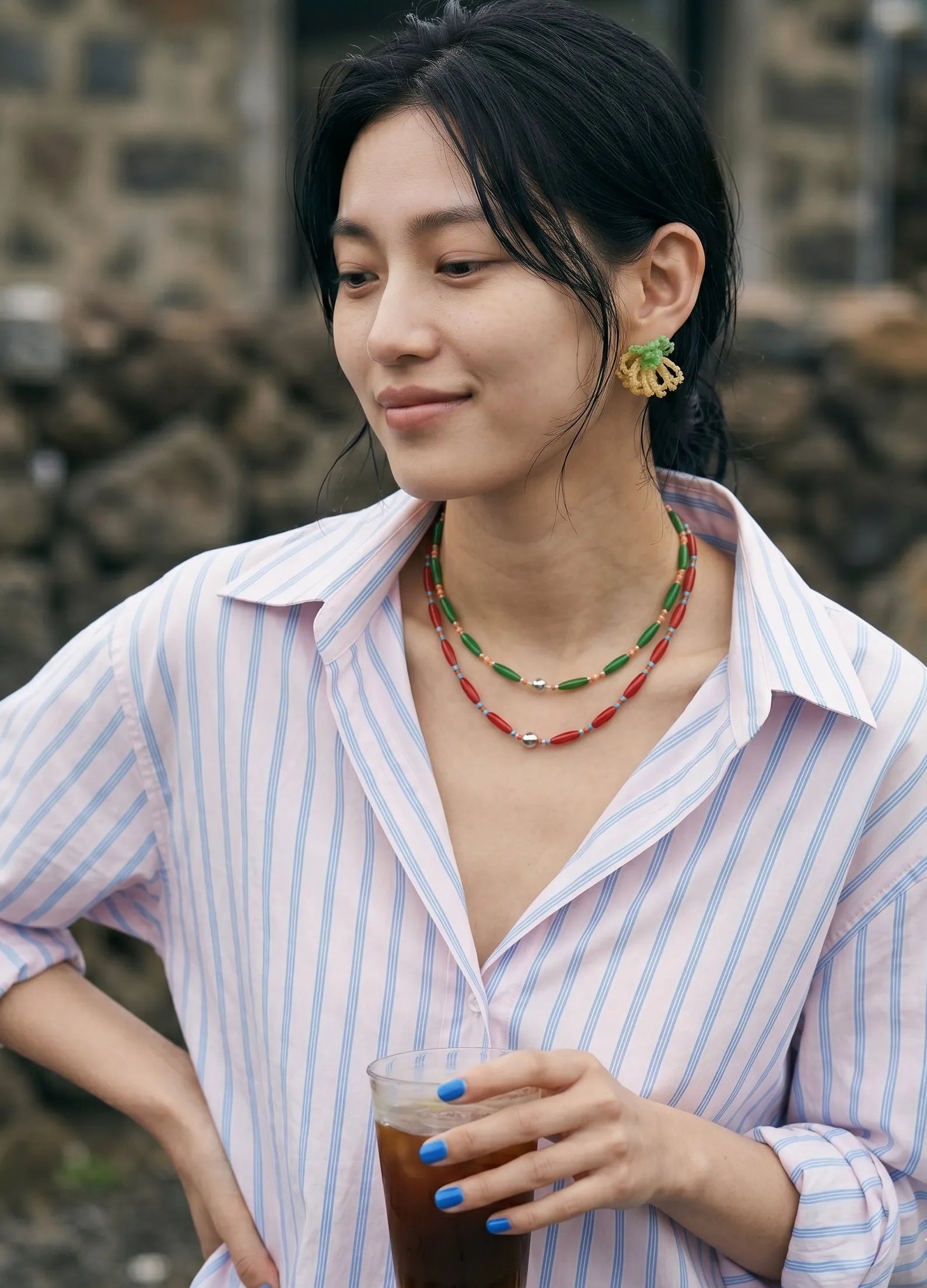 A candid lifestyle shot of a woman with a dewy, freckled makeup look holding an iced beverage. She wears an effortlessly unbuttoned striped shirt styled with mint green beaded cluster earrings and contrasting green and red layered bead necklaces