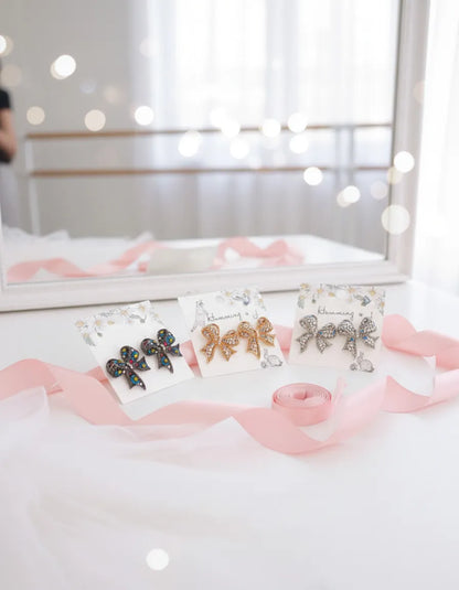 Three crystal bow stud earring colorways displayed on jewelry cards in front of a vanity mirror with pink ribbon — Antique Multi, Gold Pink, and Silver Blue — styled flatlay, handmade in Korea