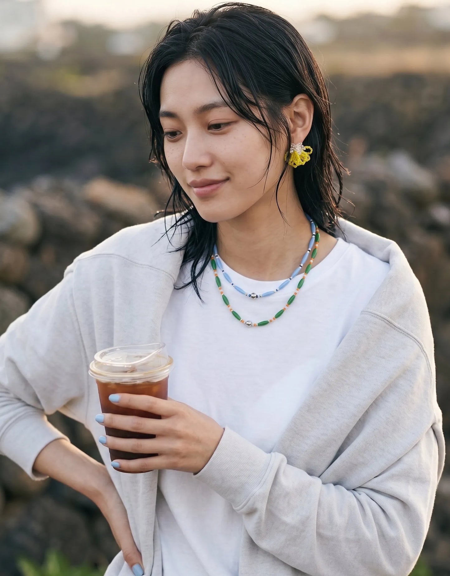 An effortless casual boho portrait featuring a woman with wet-look hair holding an iced coffee. Her minimalist outfit highlights vibrant statement yellow beaded flower earrings paired with layered multi-strand blue and green bead necklaces.