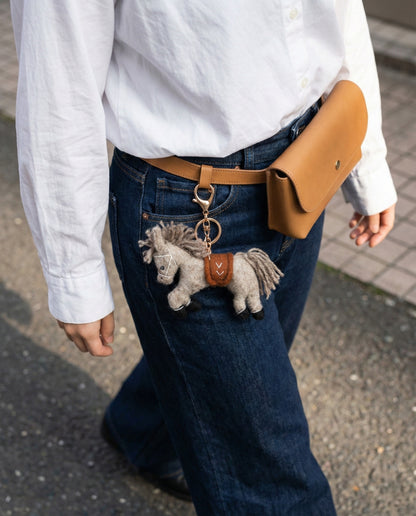 Grey wool felt pony keychain attached to camel belt bag with white linen shirt and denim jeans casual outfit
