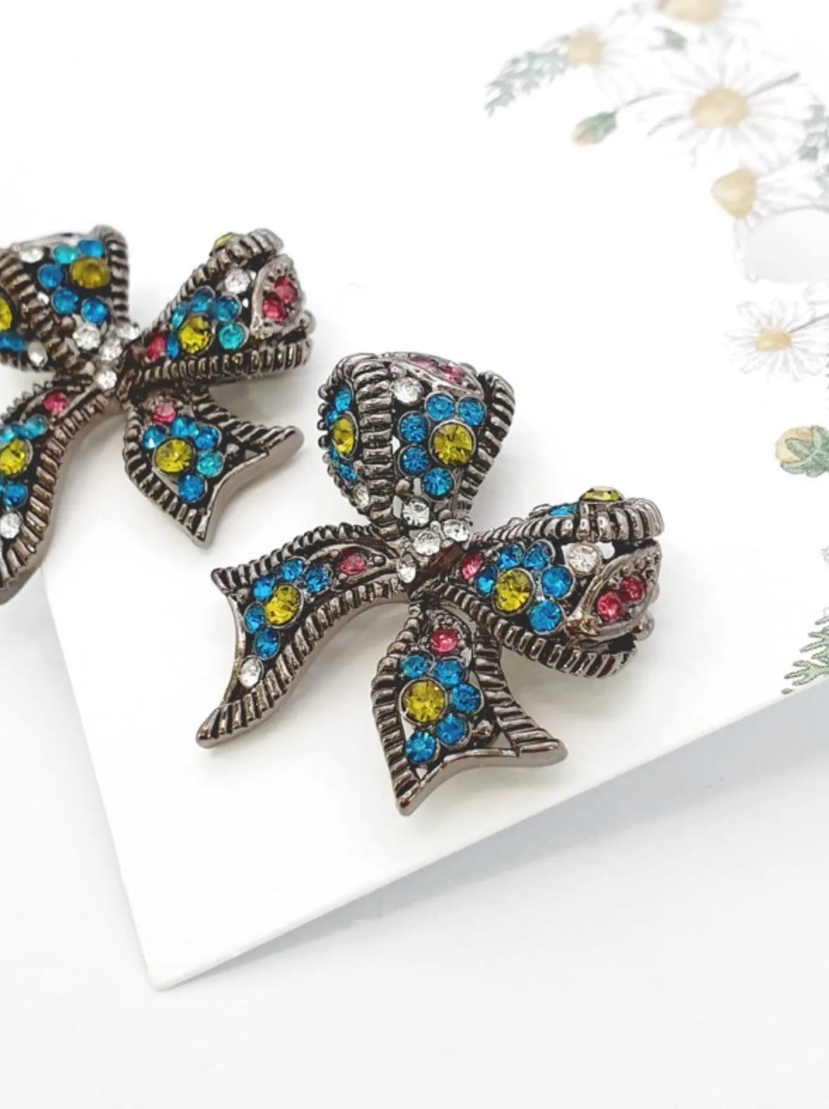 Pair of Antique Multi crystal bow stud earrings with blue, yellow, pink, and clear rhinestones on white floral background — close-up product shot, handmade in Korea