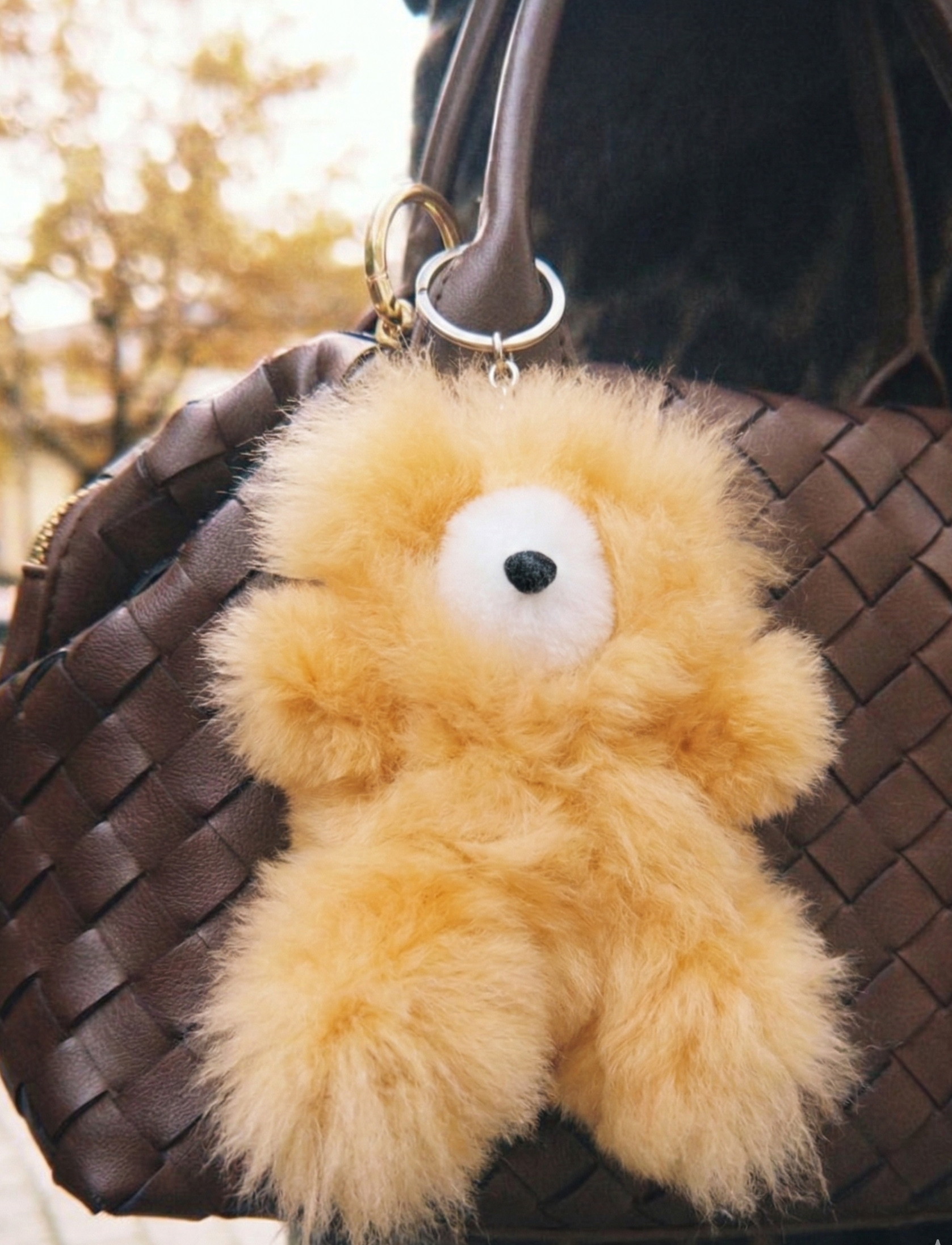 Golden yellow fluffy alpaca bear bag charm hanging from a brown woven leather backpack worn on a person, shot in warm natural outdoor light