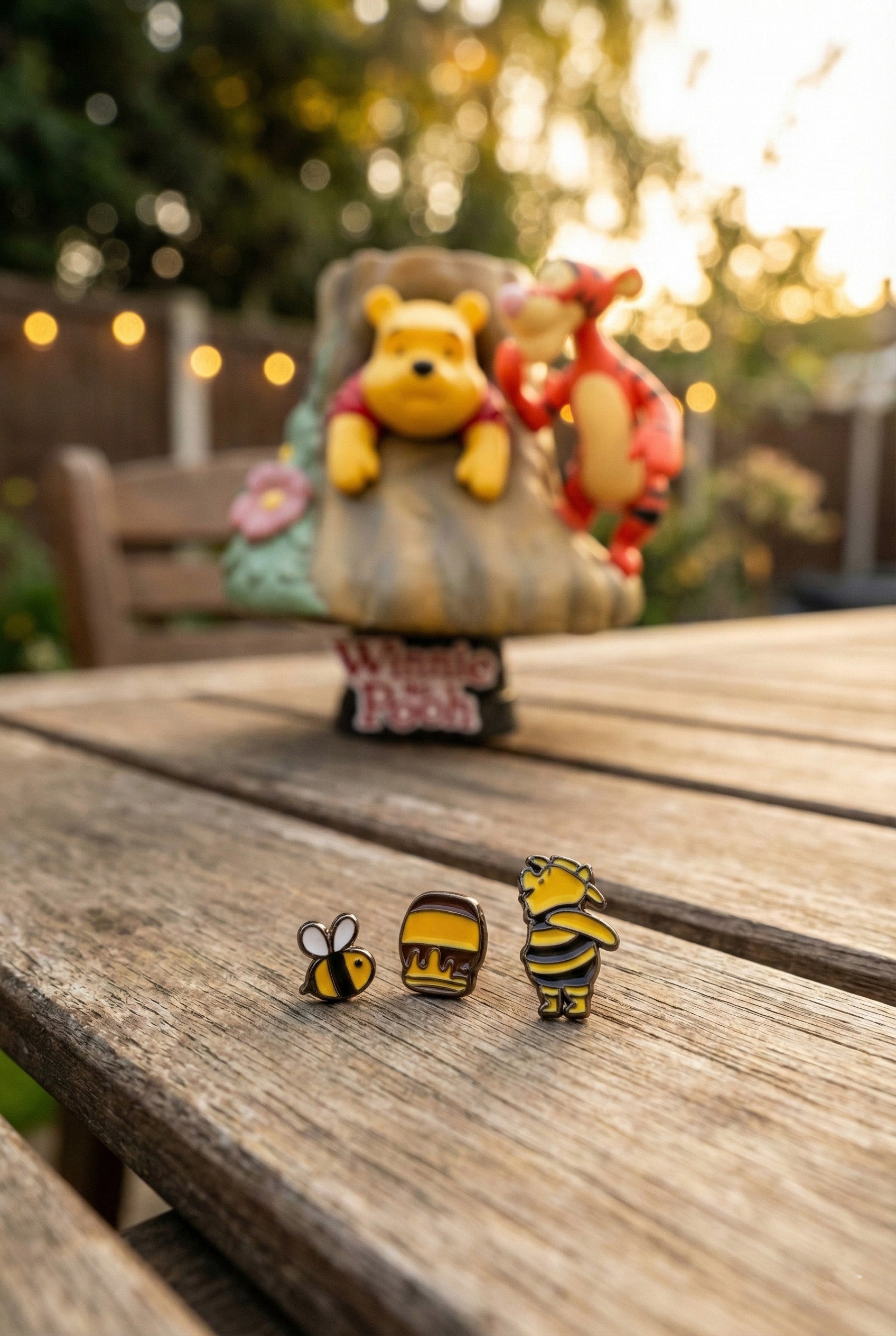 Winnie the Pooh, honey pot, and bee earrings displayed on a wooden table outdoors with a Pooh and Tigger figurine in the background.