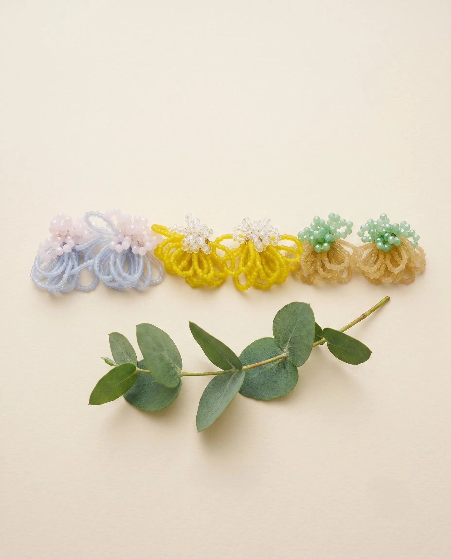 A flat lay of three pairs of handcrafted beaded cluster earrings in pastel blue, bright yellow, and mint green, placed next to a fresh eucalyptus sprig on a minimalist beige background.