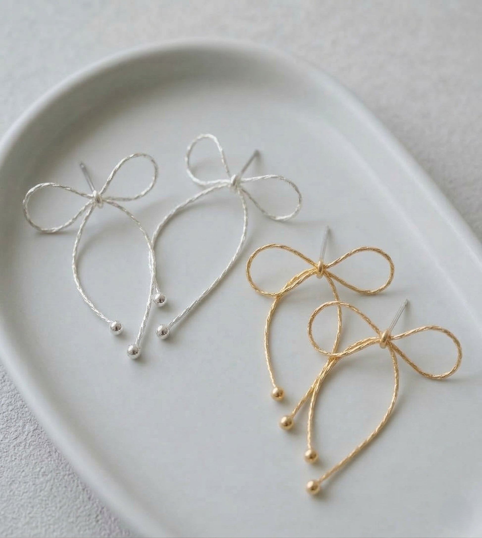 Silver and gold textured chain bow drop earrings displayed on a white ceramic plate showing both color options side by side