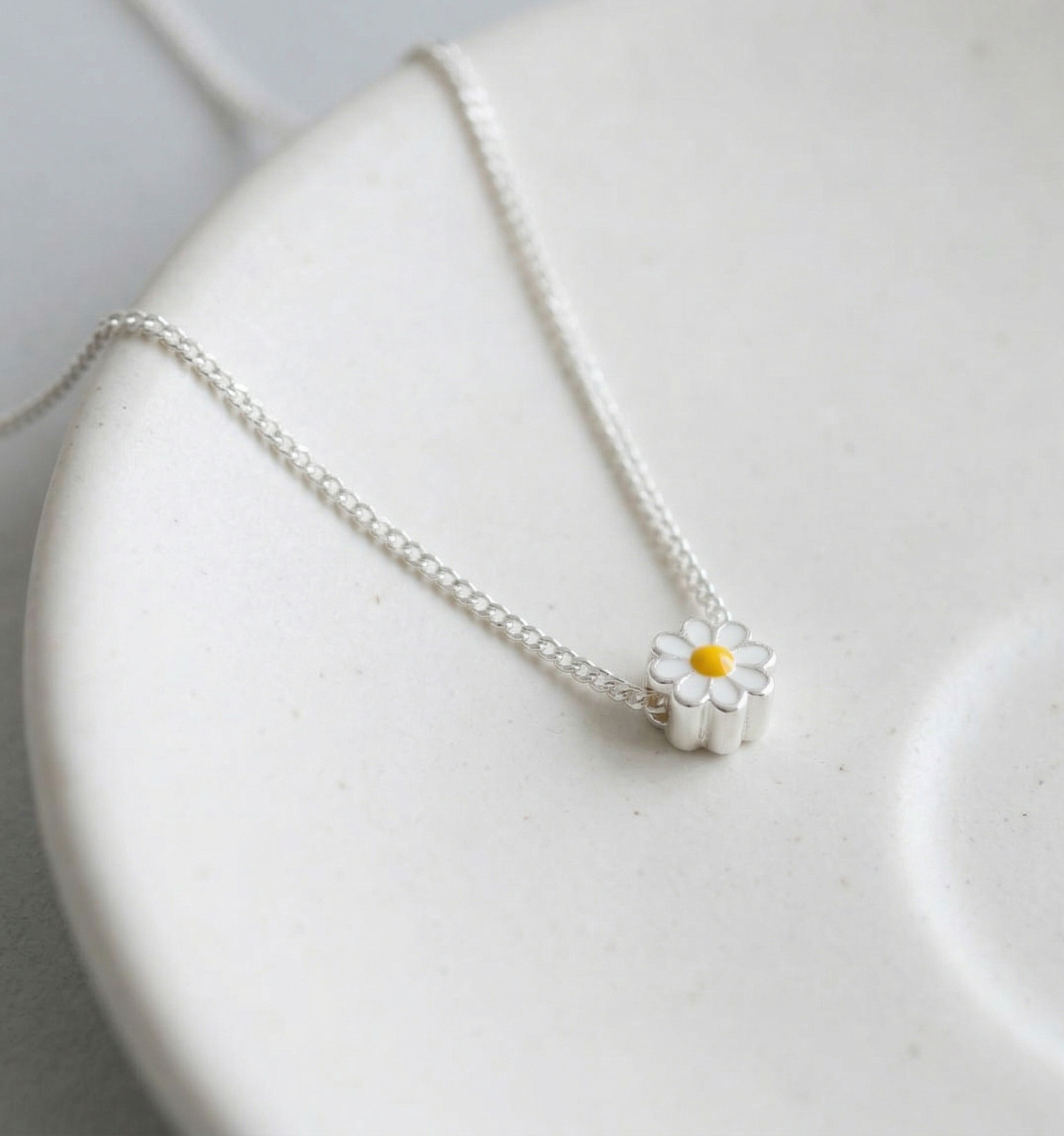 Dainty 925 sterling silver daisy flower pendant necklace with white enamel petals and yellow enamel center, displayed on a white ceramic plate