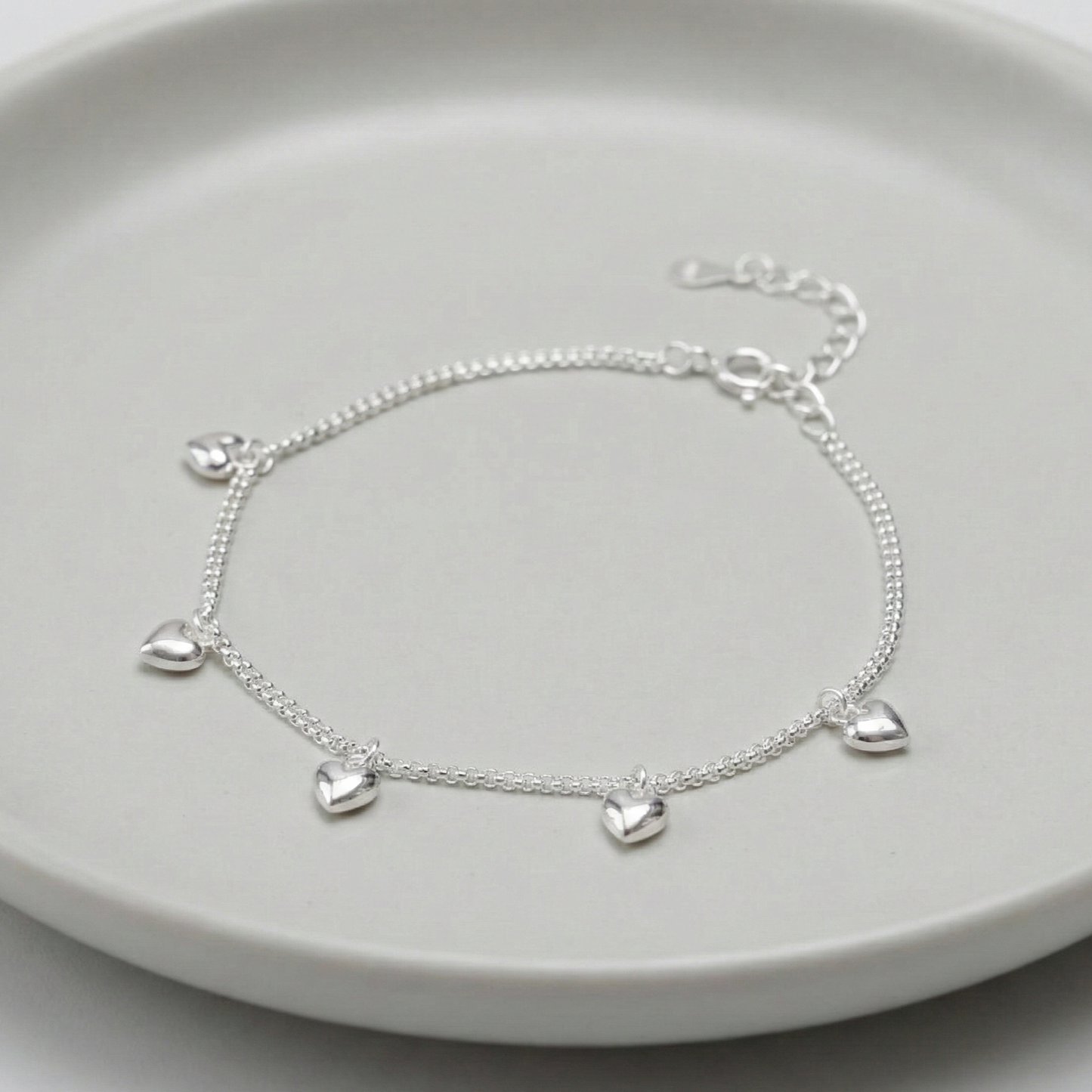 925 sterling silver double chain puffed heart charm bracelet laid on a grey ceramic plate, showing 5 dangling 3D heart charms and adjustable extender chain