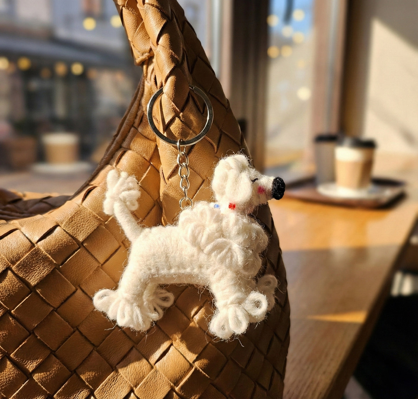 Handmade wool felt poodle keychain hanging on a woven brown leather bag in a cozy cafe setting