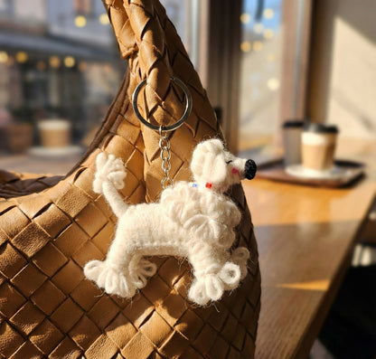 Handmade wool felt poodle keychain hanging on a woven brown leather bag in a cozy cafe setting