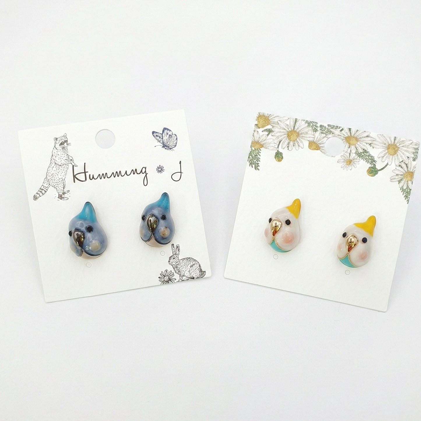 Two pairs of handmade bird stud earrings on illustrated earring cards — cockatiel and blue parrot 