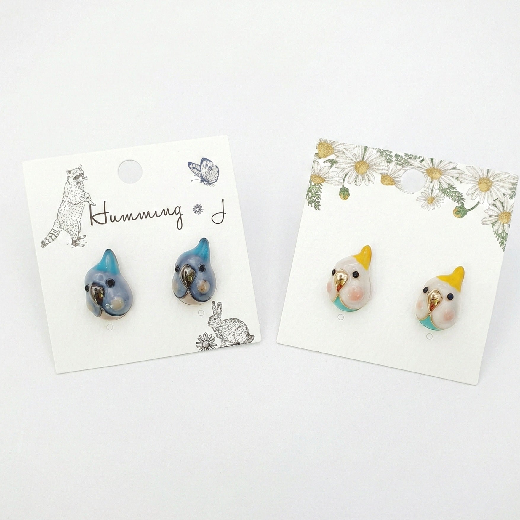 Two pairs of handmade bird stud earrings on illustrated earring cards — cockatiel and blue parrot 
