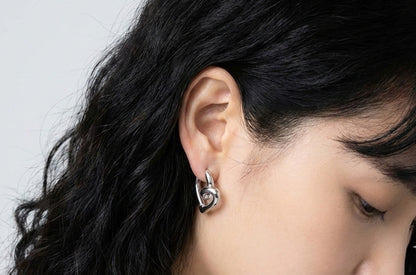 Woman wearing silver twisted heart hoop earrings, sculptural love knot huggie design with titanium posts