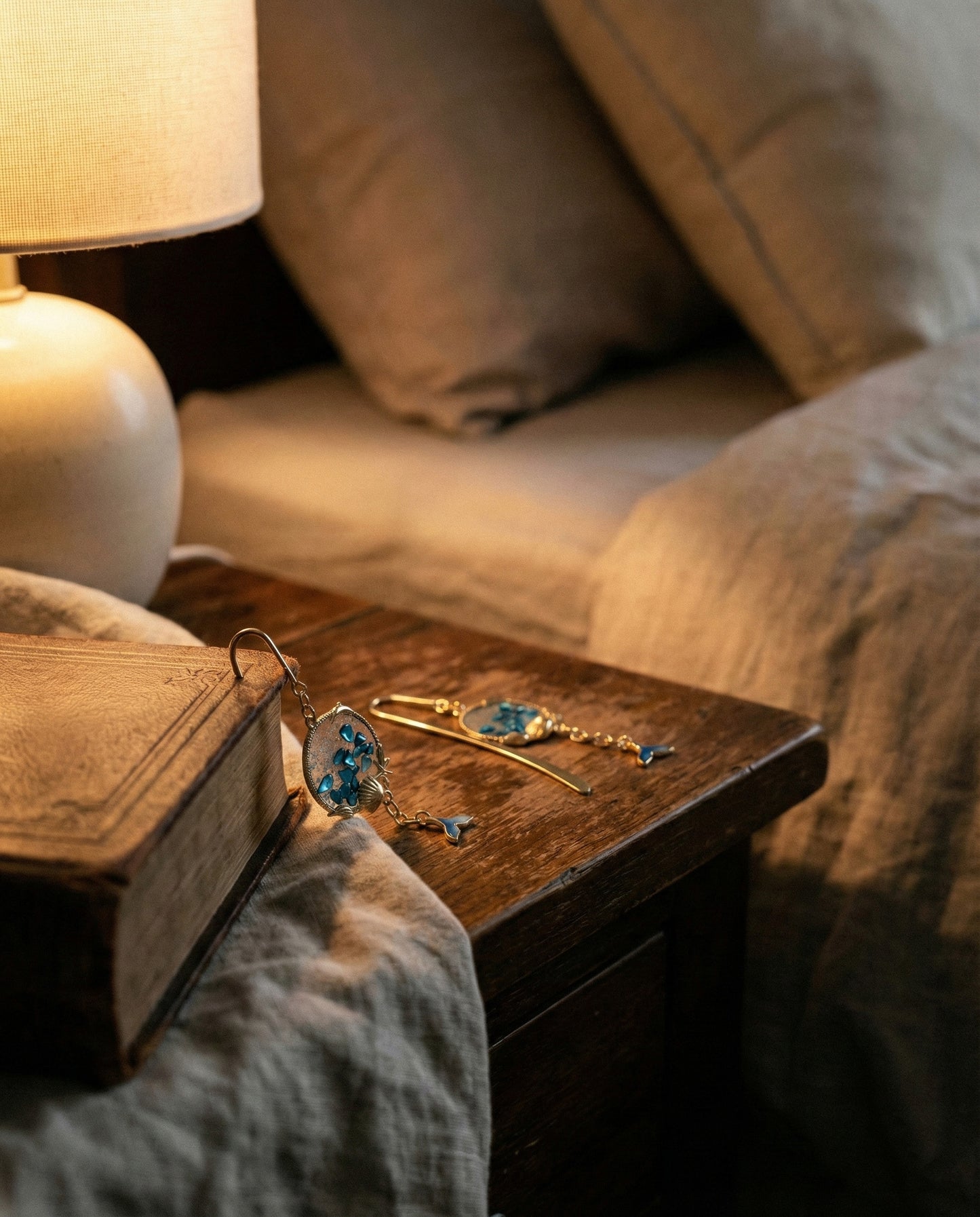 Whale tail metal bookmark with ocean blue resin charm on nightstand with book and warm lamp light