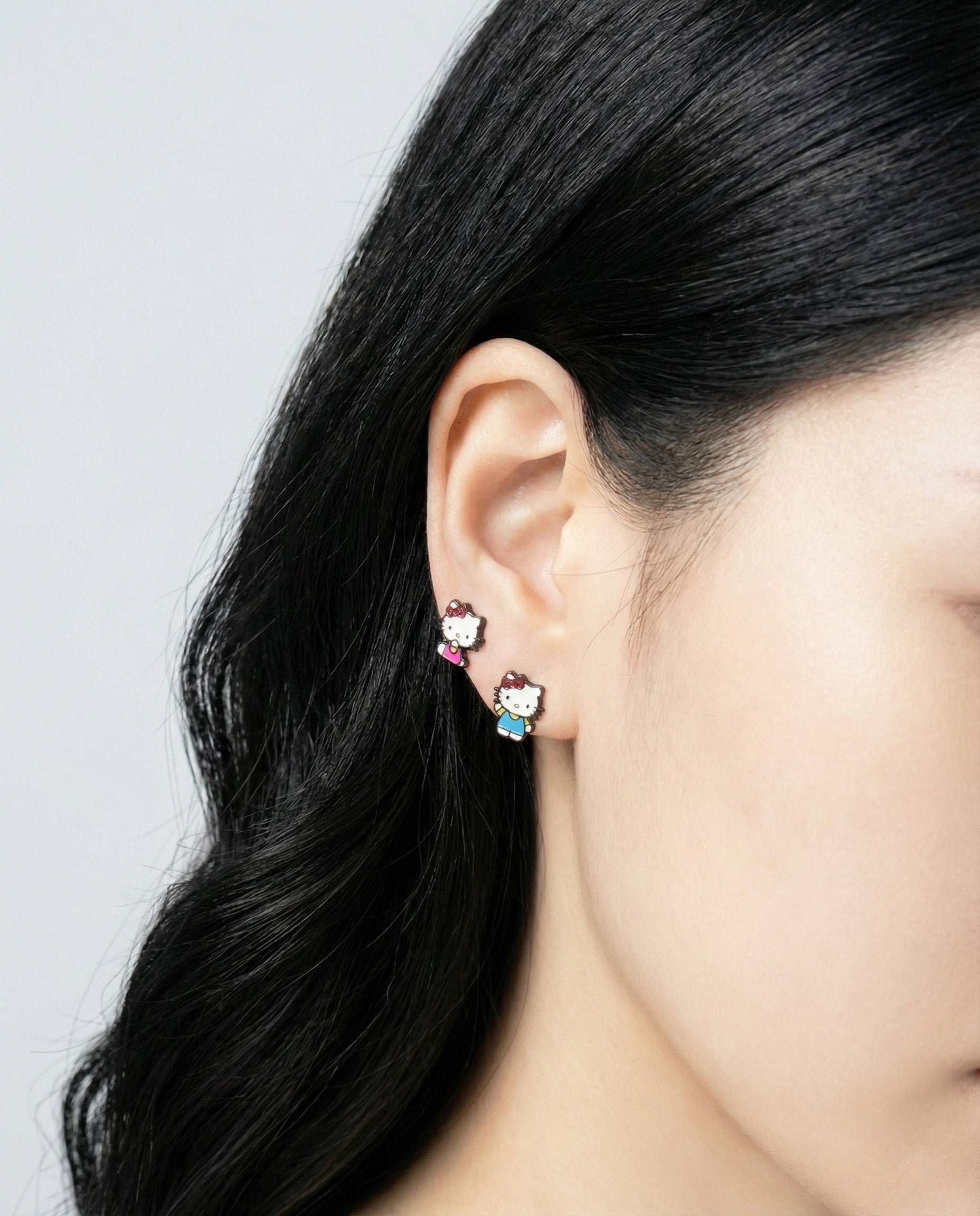 Close-up of model wearing Hello Kitty asymmetrical stud earrings showing blue overalls and pink ribbon designs