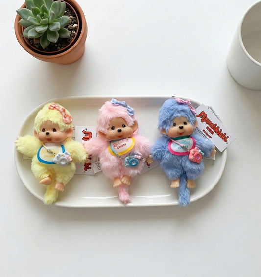 Monchhichi camera plush keyring in three pastel colors - yellow, pink, and blue on white ceramic plate