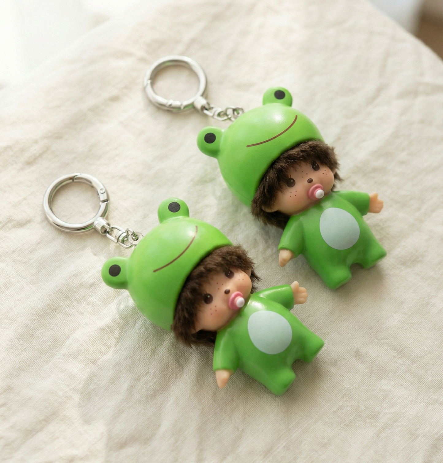 Two green frog baby doll keychains on linen fabric showing size comparison and freckle pacifier detail