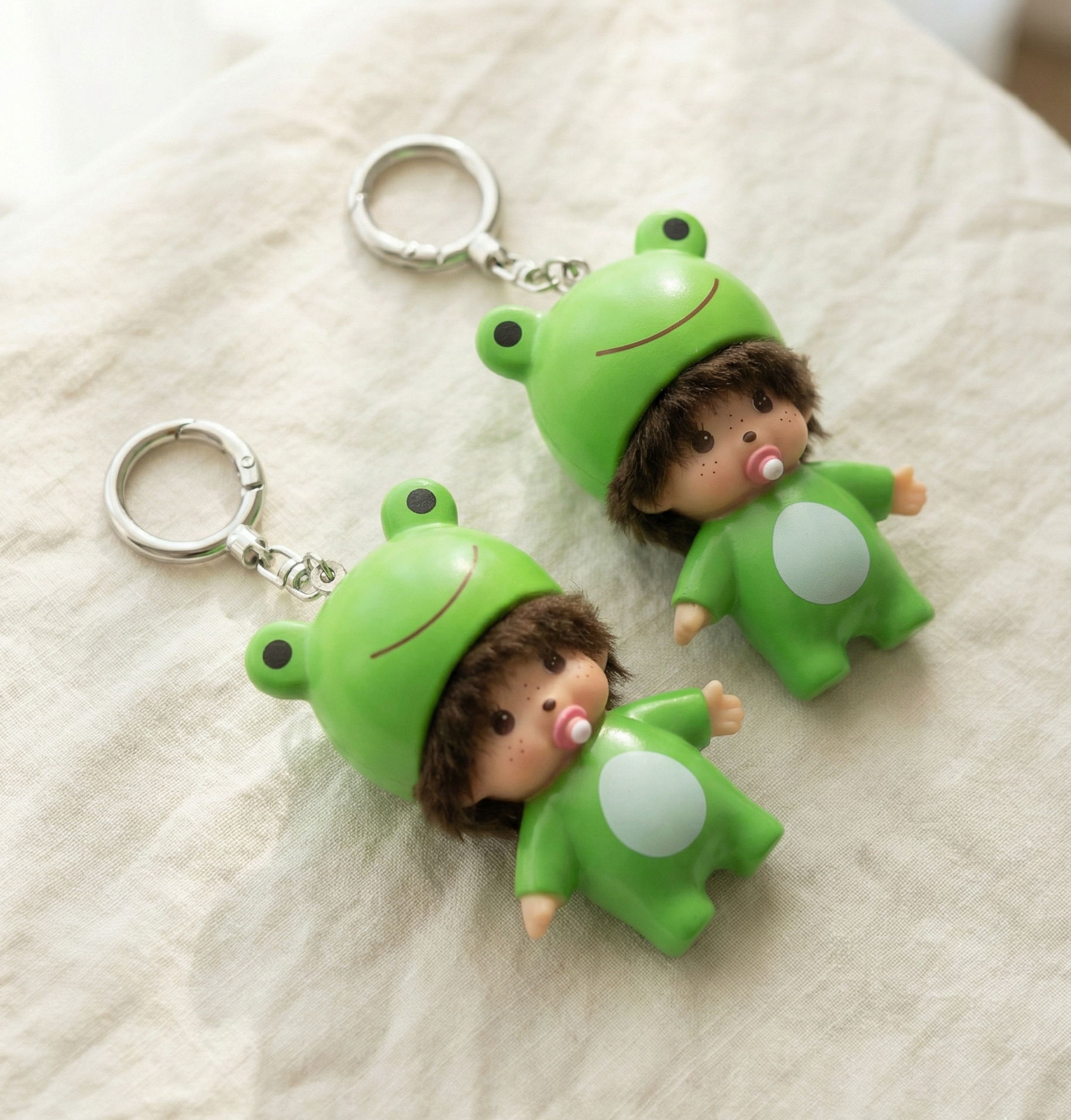 Two green frog baby doll keychains on linen fabric showing size comparison and freckle pacifier detail