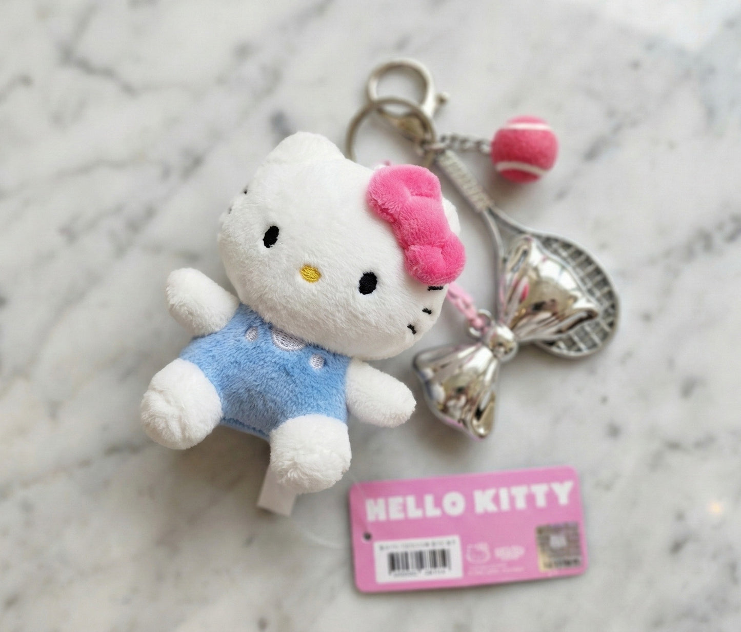 Authentic Hello Kitty plush keychain with silver tennis racket, pink tennis ball, and silver bow charm on white marble background
