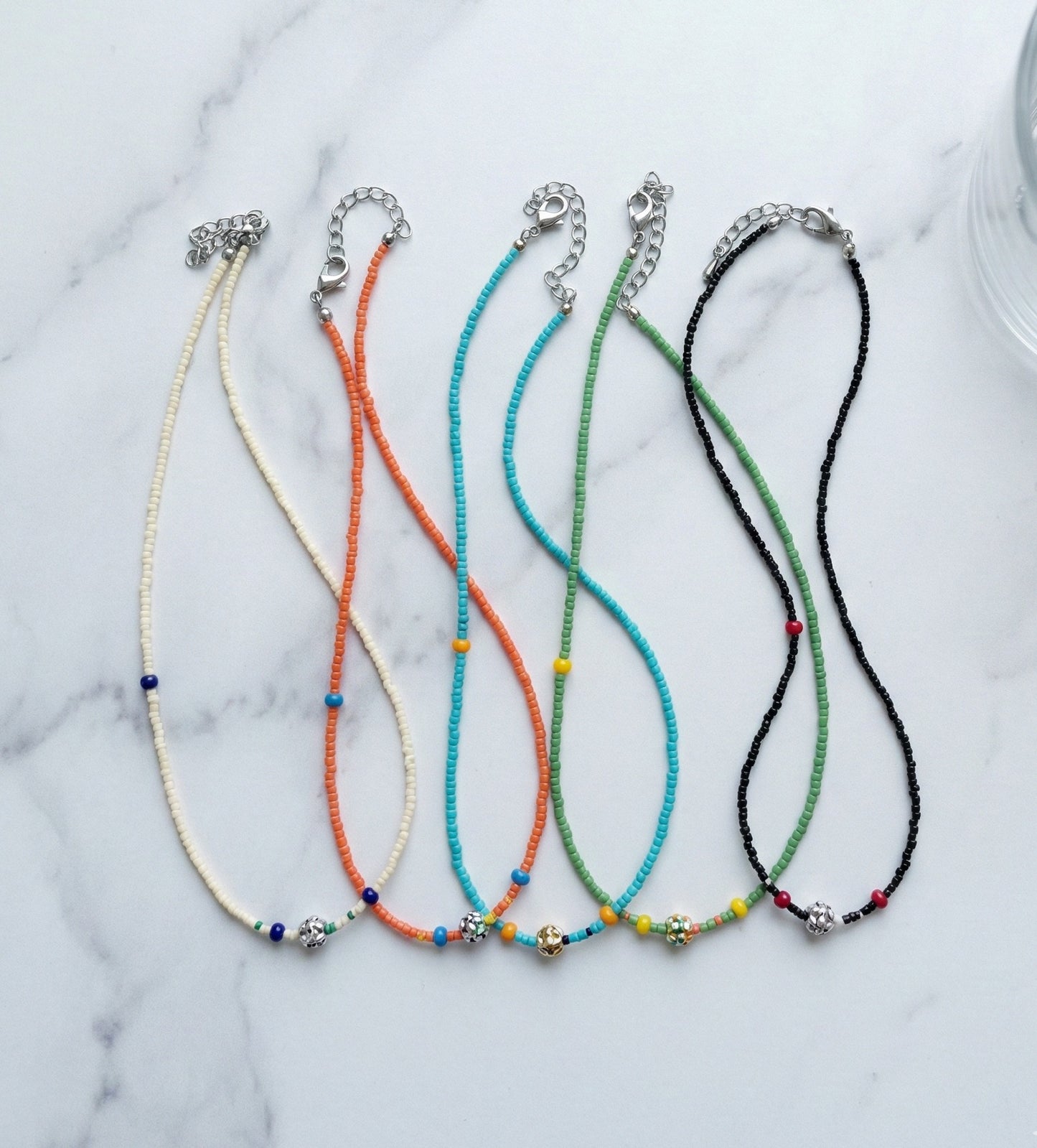 Five colorful four-leaf clover seed bead choker necklaces in ivory, black, turquoise, orange, and green arranged on white marble
