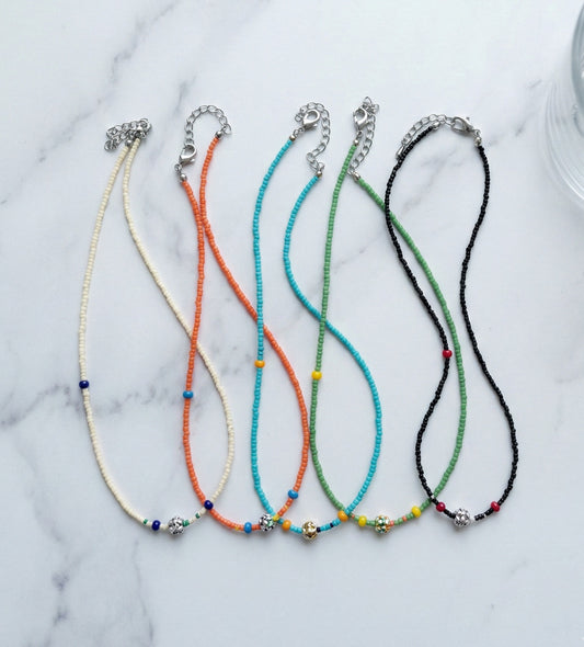 Five colorful four-leaf clover seed bead choker necklaces in ivory, black, turquoise, orange, and green arranged on white marble